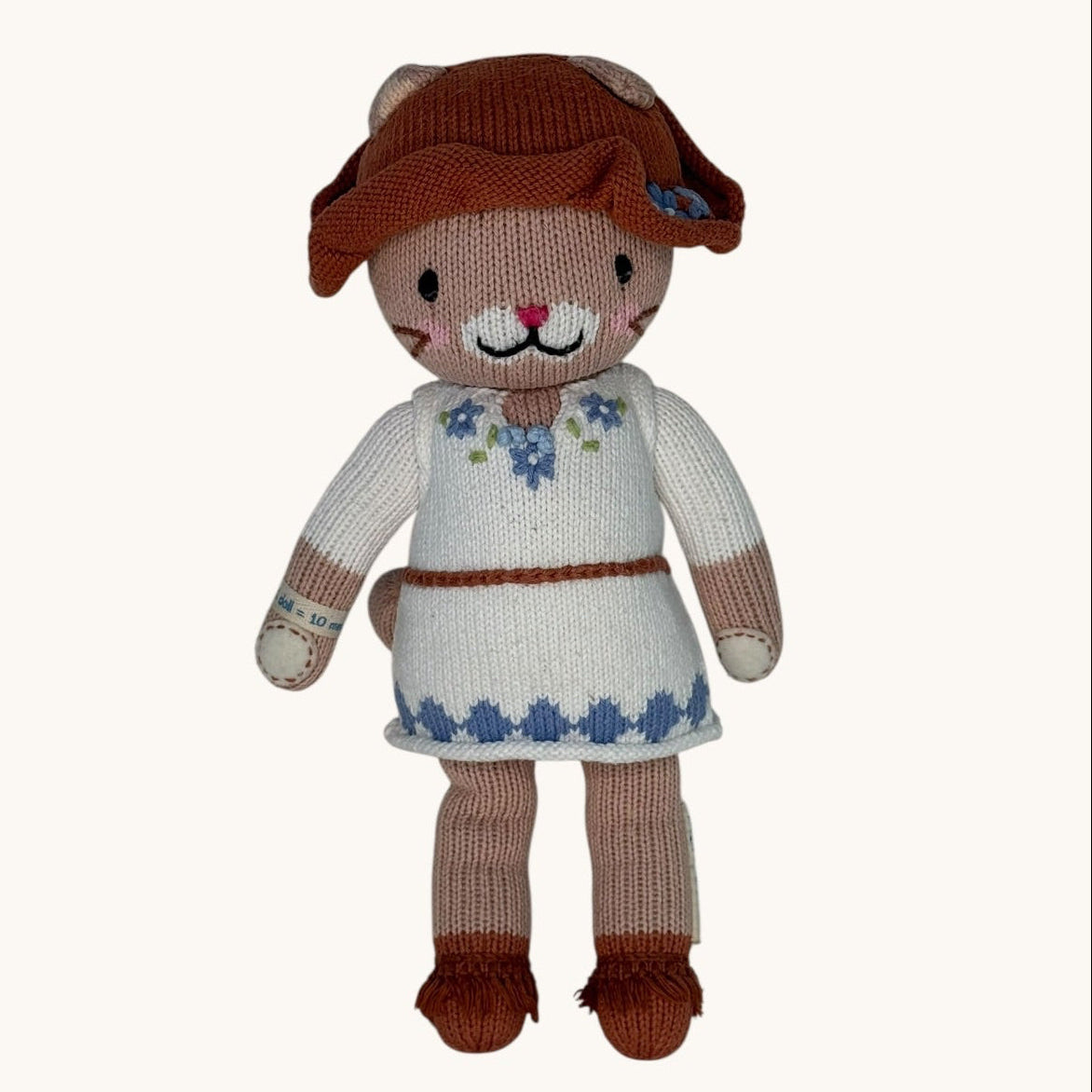 Knitted toy cat wearing a brown hat and white dress with blue patterns on a white background