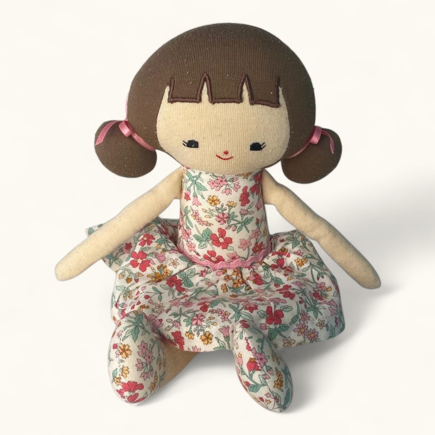 Stuffed doll with brown hair, floral dress, and pink buns on a white background