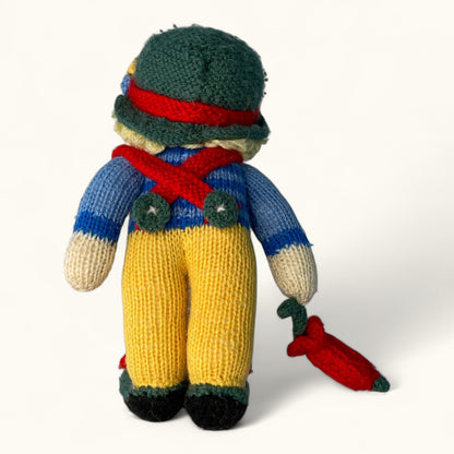 Knitted toy figure with a green hat, blue sweater, yellow pants, and red scarf on a white background.