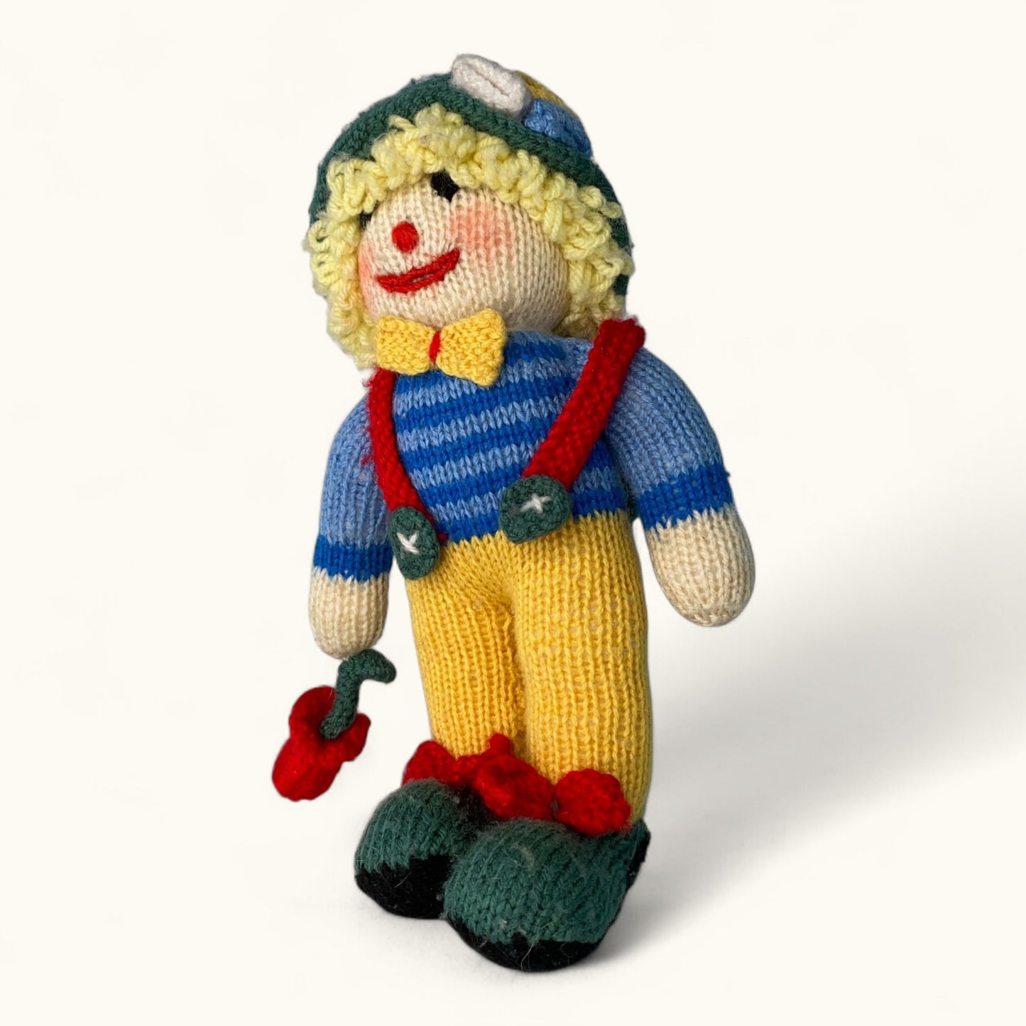 Knitted toy figure of a person wearing a blue sweater, yellow pants, and a green hat on a white background.