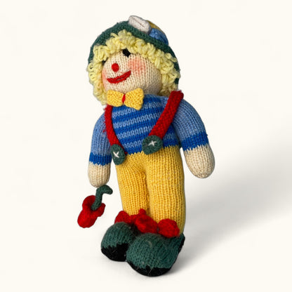 Knitted toy figure of a person wearing a blue sweater, yellow pants, and a green hat on a white background.