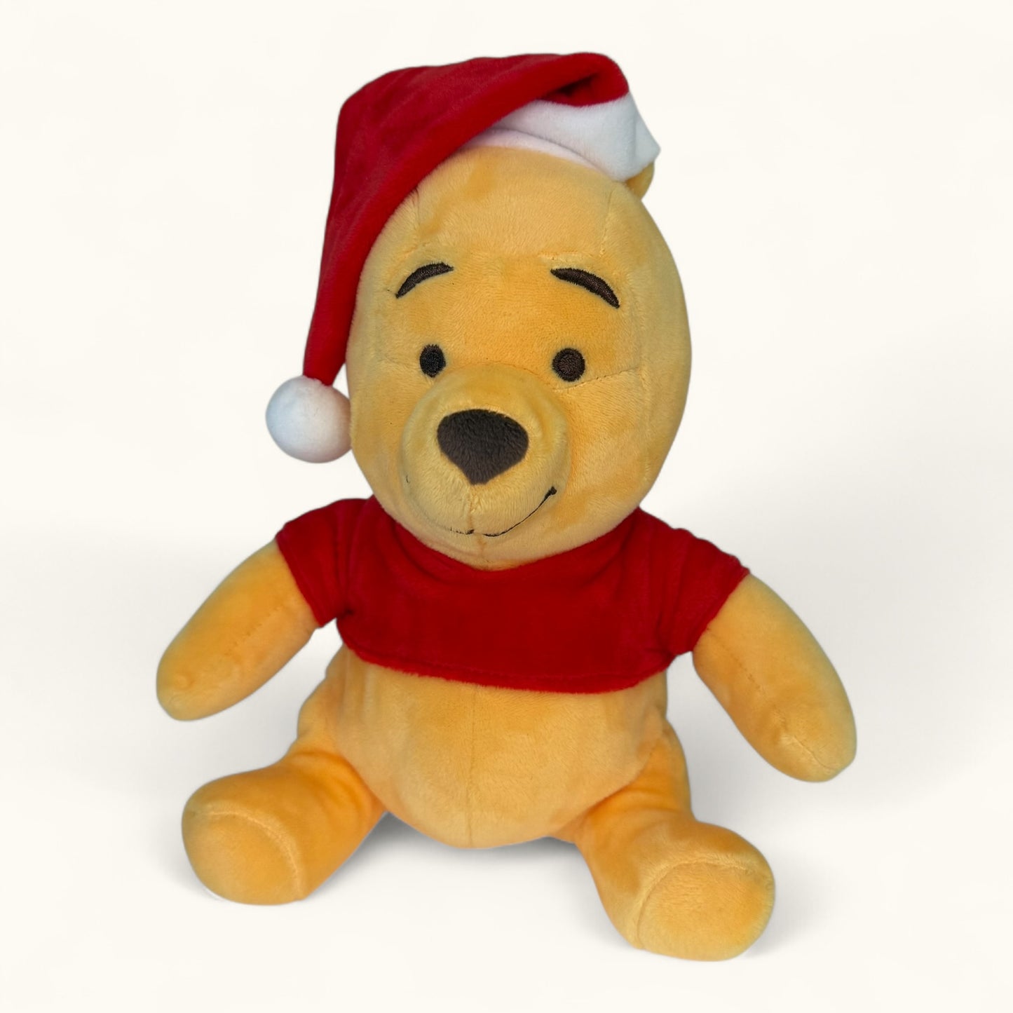 Plush toy Winnie the Pooh bear wearing a red shirt and Santa hat on a white background
